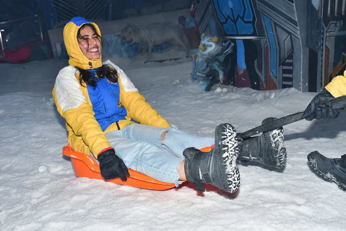 A young woman in a colorful jacket laughs while sledding on snow. She is wearing ripped jeans and black boots.