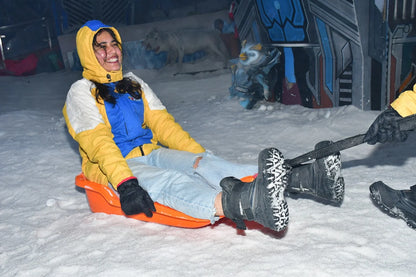 A young woman in a colorful jacket laughs while sledding on snow. She is wearing ripped jeans and black boots.