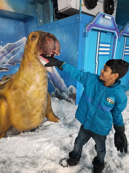 A young boy in a blue jacket playfully feeds a large seal statue in a snowy, indoor attraction.