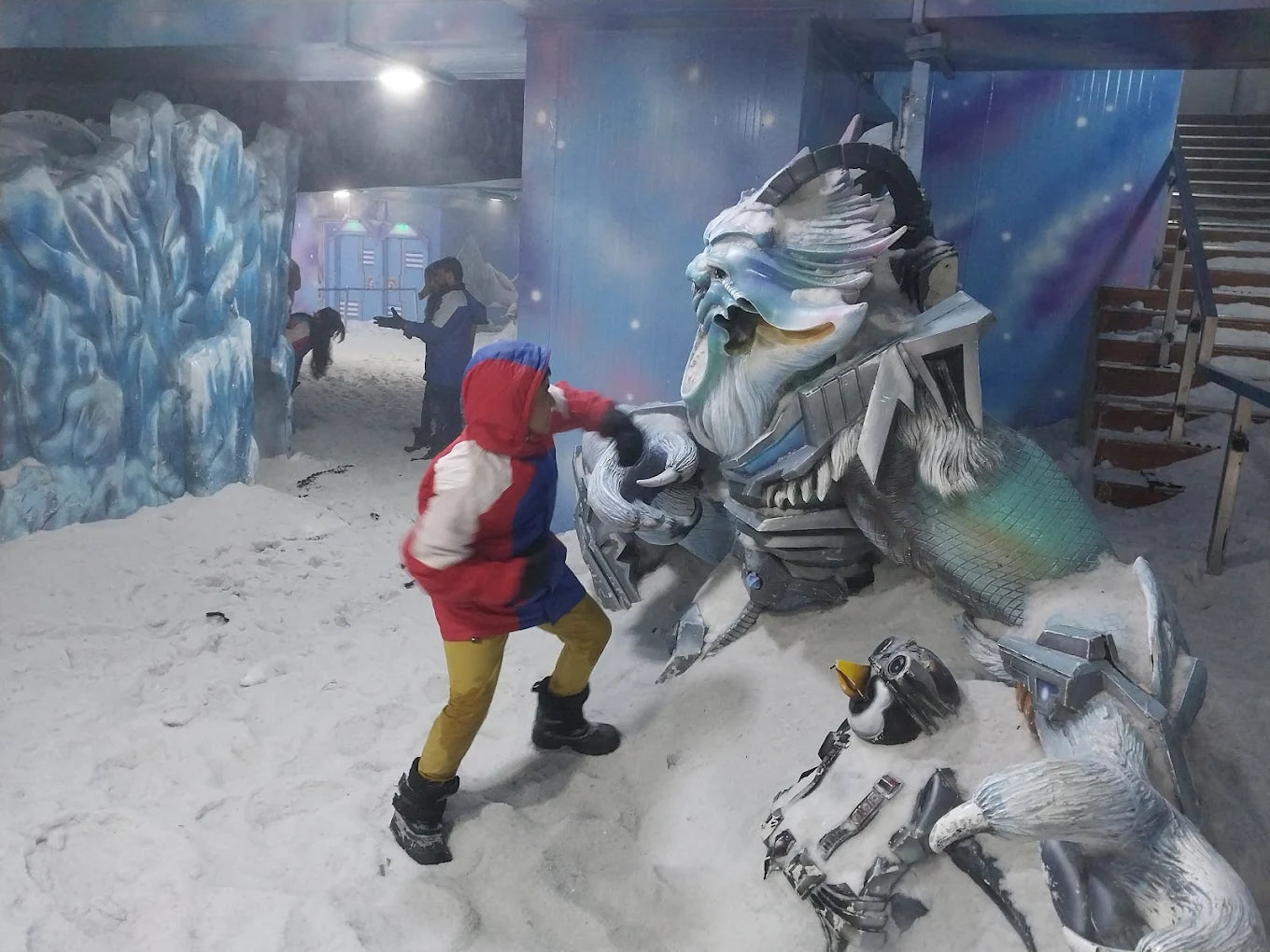 A child in a red, white, and blue jacket plays in a snowy indoor park with a large ice monster statue.