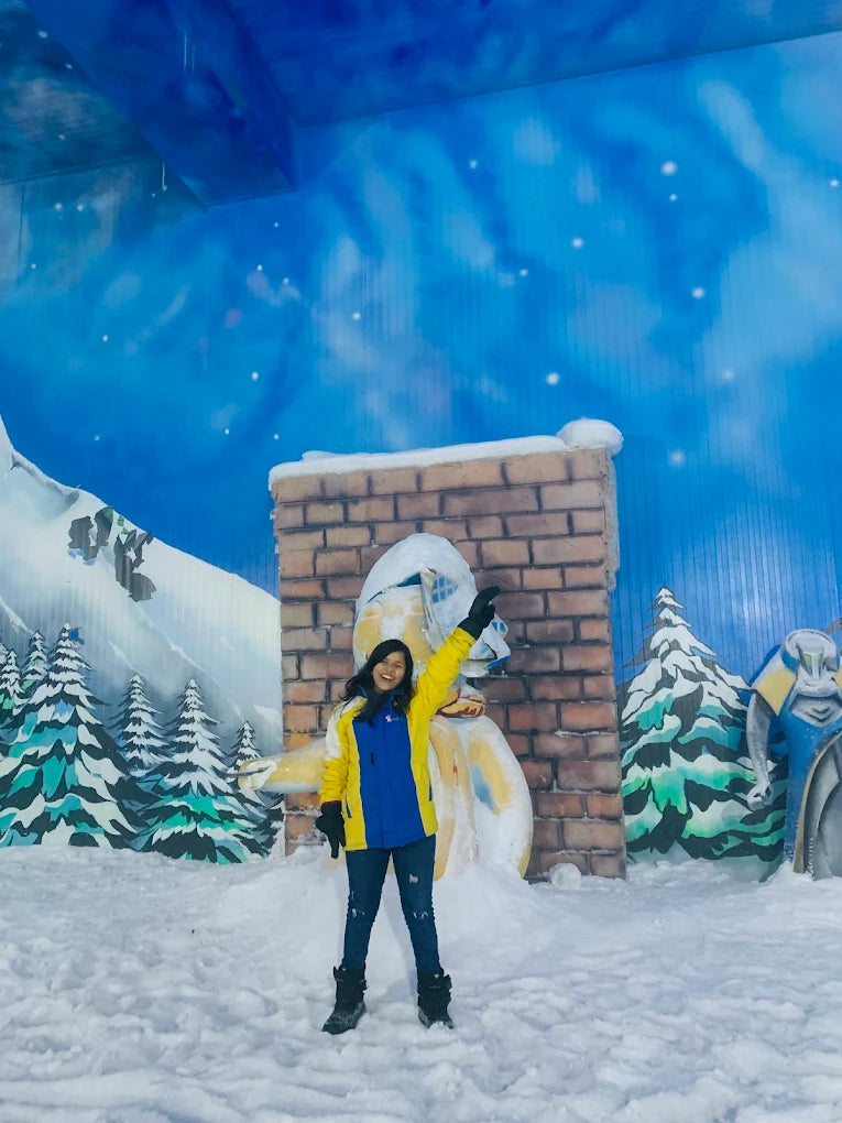 A young woman in a yellow and blue jacket poses in a snowy indoor winter wonderland.