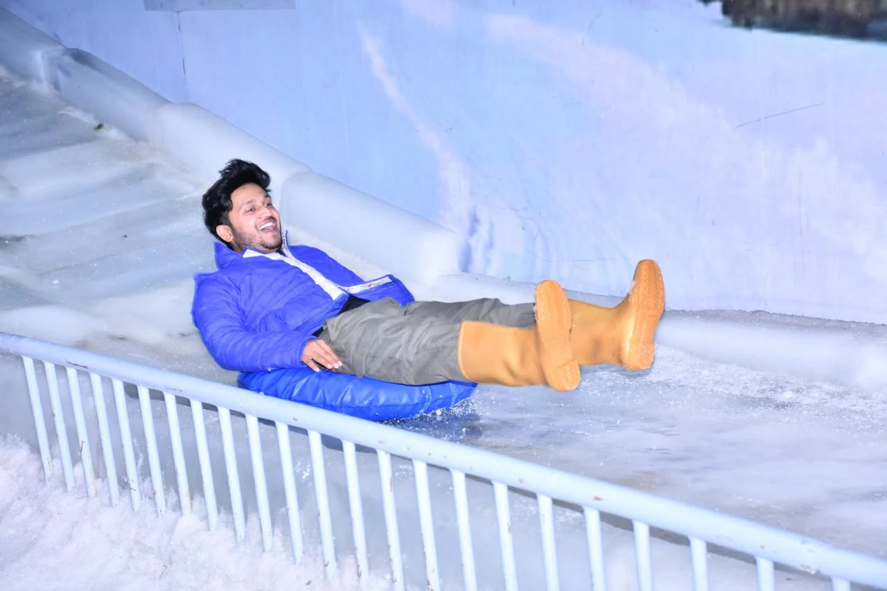 A young man slides down an ice slide on a blue tube, wearing a blue jacket and tan boots.