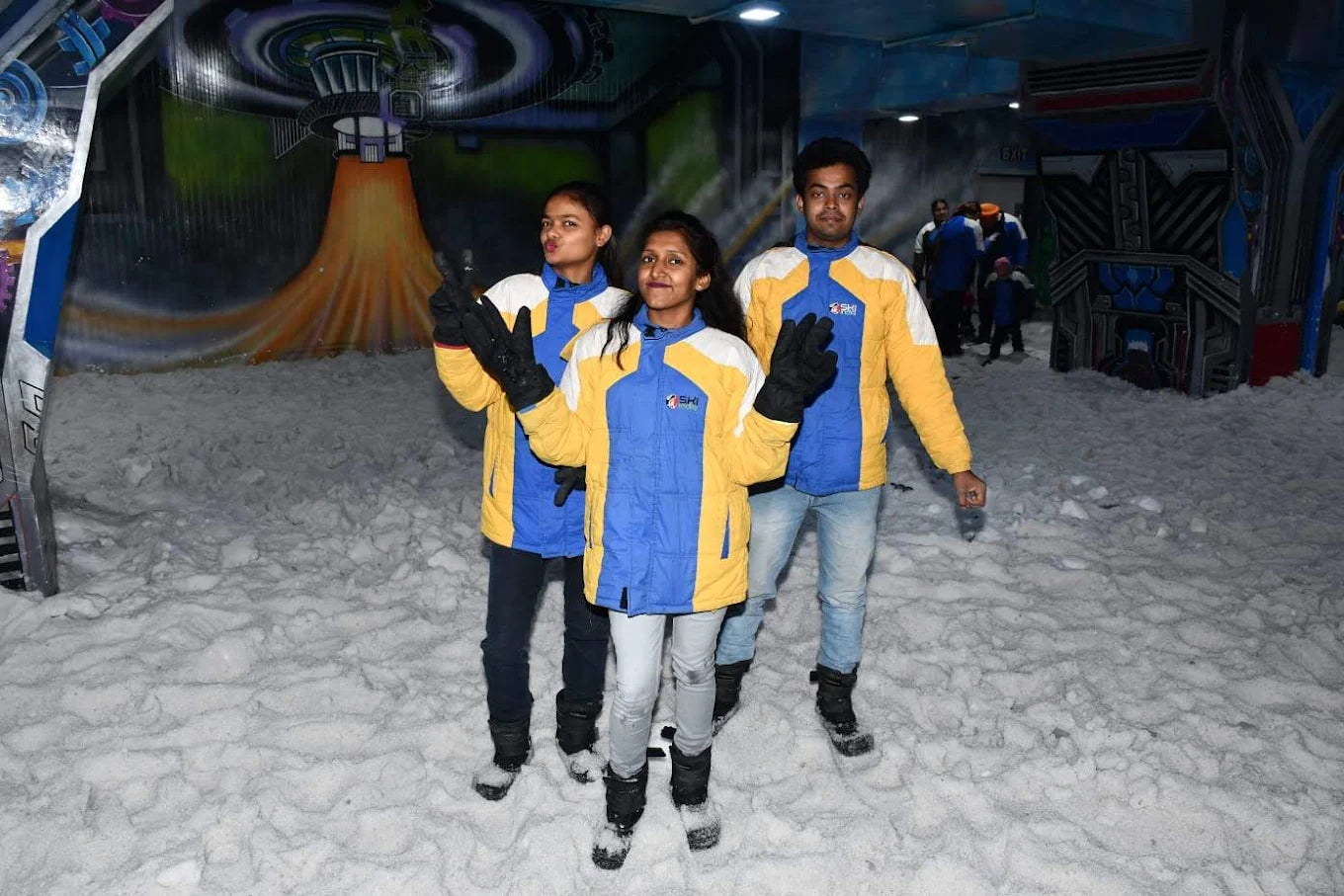 Three young people in ski jackets pose in a snowy indoor park with a mural.