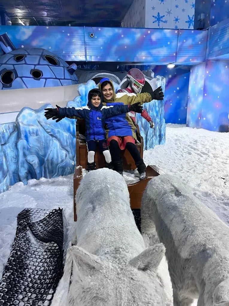 Two children in a sleigh with a person in a robot costume in a snowy indoor park.