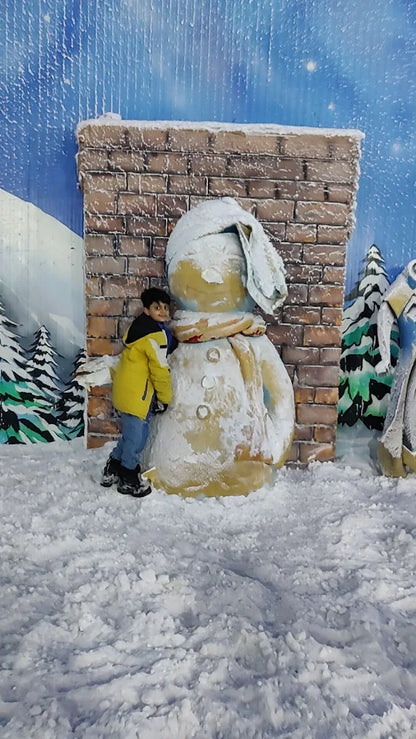 A young boy in a yellow jacket hugs a large snowman in a snowy, winter-themed setting.