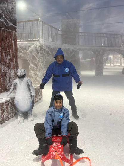 A young boy on a sled is pushed by an adult in a snowy indoor park.