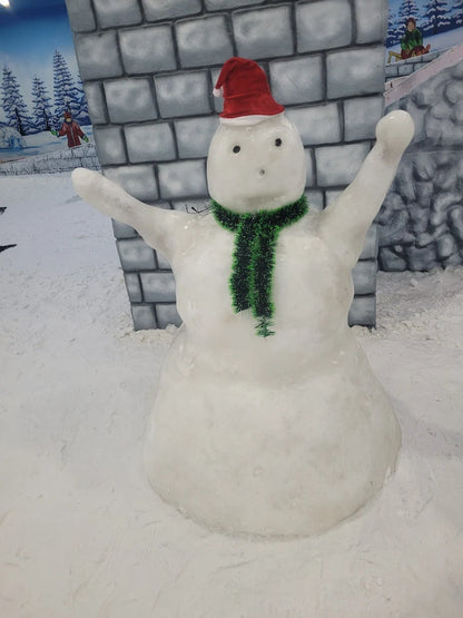 A snowman with a Santa hat and green scarf stands in a snowy area with brick-like walls and painted trees.