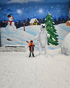 A child poses with a polar bear mascot in a snowy, indoor winter wonderland with a snowman and a Christmas tree.
