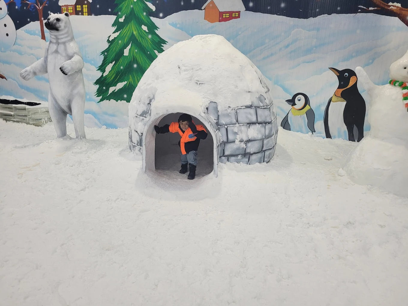 A child in an orange jacket peeks out of an igloo in a snowy, arctic-themed attraction with polar bears and penguins.