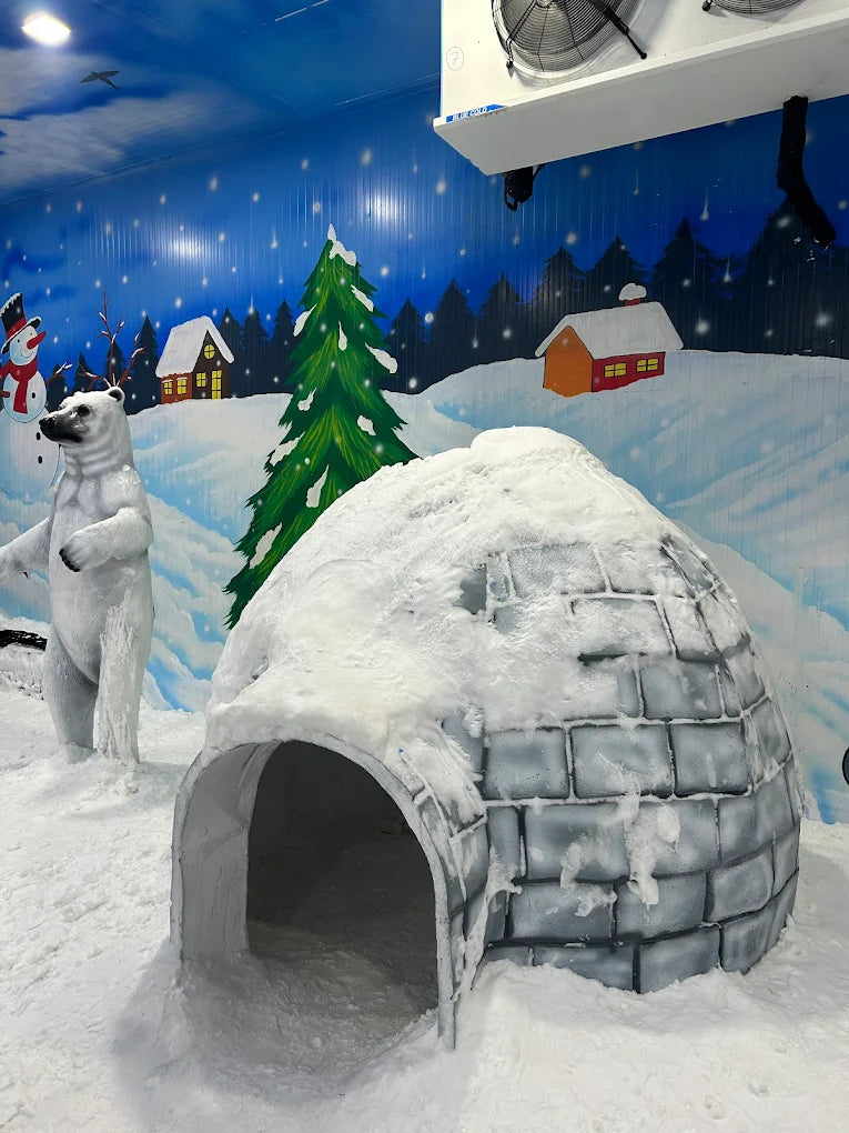 A polar bear stands in front of an igloo in a snowy, winter-themed indoor attraction.