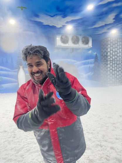 A young man in a red and gray snowsuit smiles and waves in a snowy indoor environment with a winter mural.