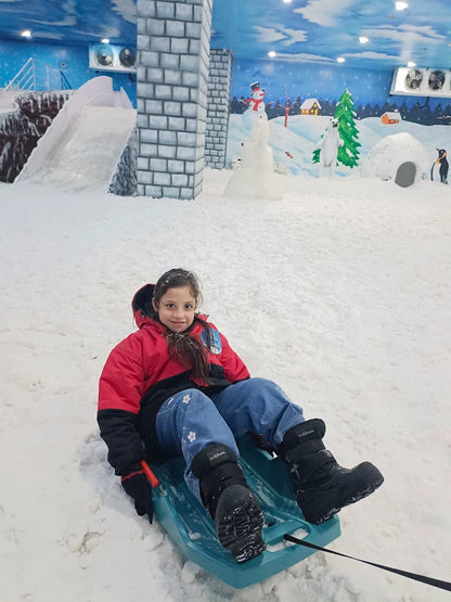 A young girl in a red jacket and blue jeans sits on a sled in a snowy indoor park. She is smiling at the camera.