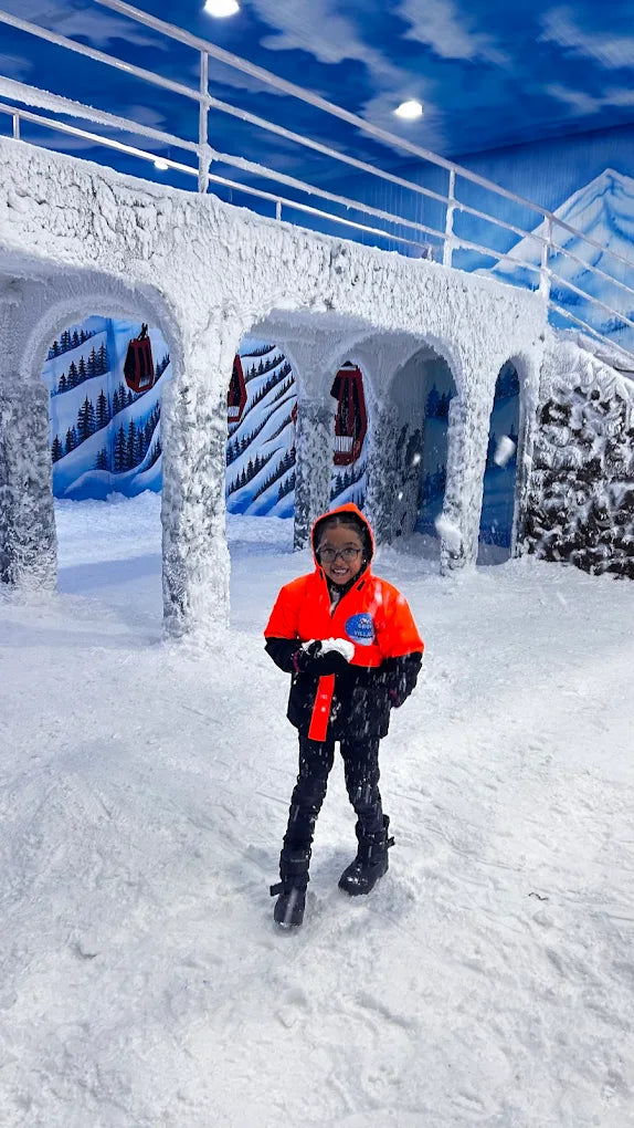 A young person in a snowsuit smiles in a snowy, indoor winter wonderland.