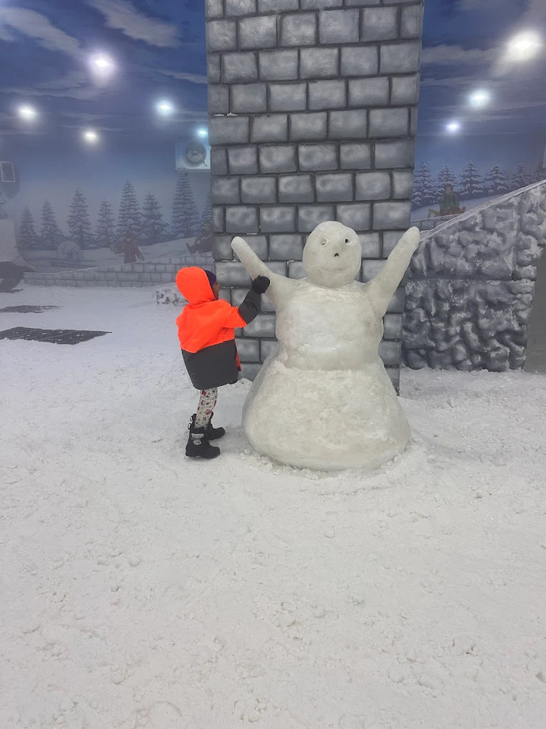 A child in an orange jacket builds a snowman in a snowy indoor park.