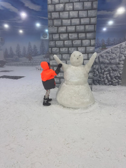 A child in an orange jacket builds a snowman in a snowy indoor park.