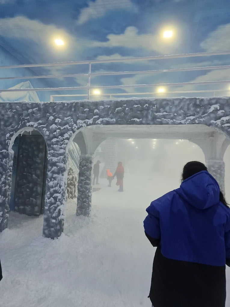 A woman in a blue jacket walks through a snowy indoor attraction with faux ice structures and a cloudy sky ceiling.