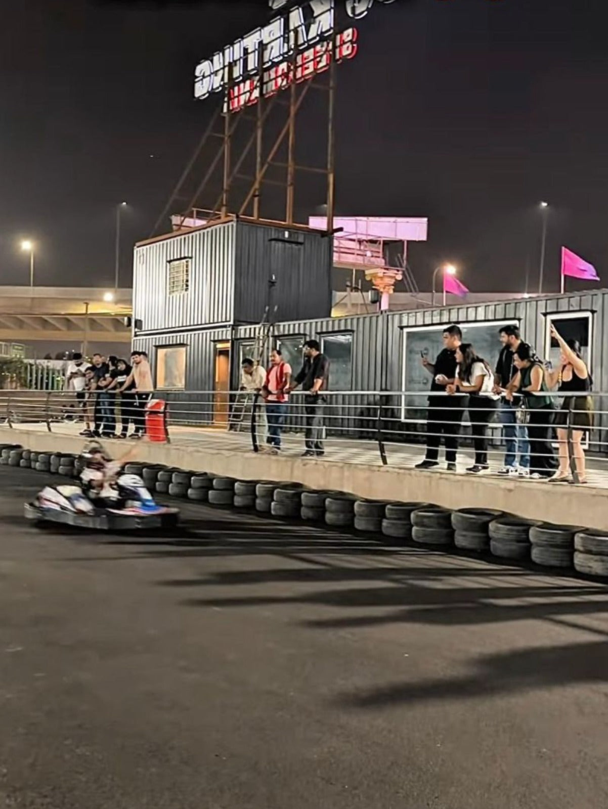 Young adults watch a go-kart race at night, with shipping containers and a large sign in the background.
