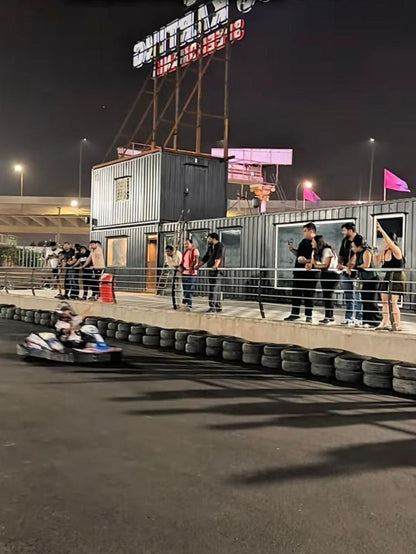 Young adults watch a go-kart race at night, with shipping containers and a large sign in the background.