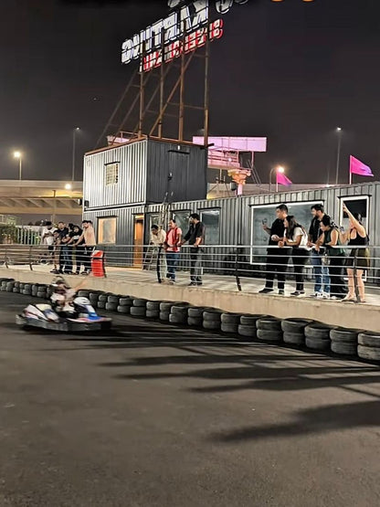 Adults watch a go-kart race at night from a raised platform overlooking the track.