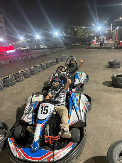 Two children in go-karts at a nighttime track, one giving a thumbs up.