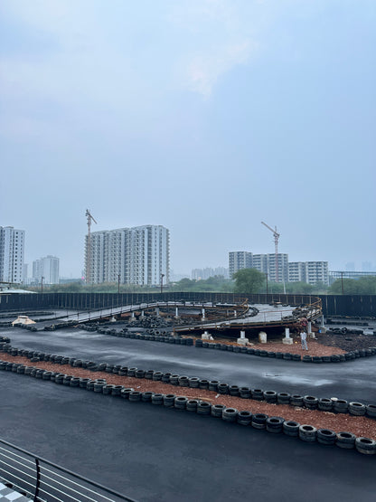 A go-kart track with tire barriers and elevated sections, under a cloudy sky with construction cranes and buildings in the background.