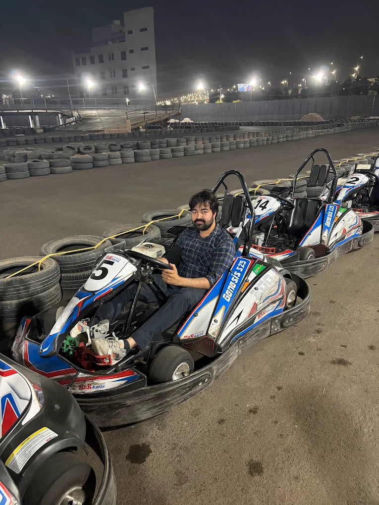 A young man sits in a go-kart at an outdoor track at night. He is smiling at the camera.