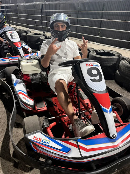 A young man in a helmet sits in a go-kart, making a hand gesture. He is at a go-kart track.