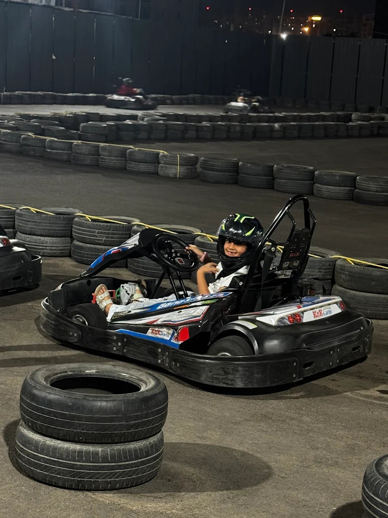 A child in a helmet smiles while sitting in a go-kart on a track at night. The track is lined with tires.
