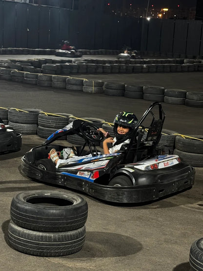 A child in a helmet smiles while sitting in a go-kart on a track at night. The track is lined with tires.