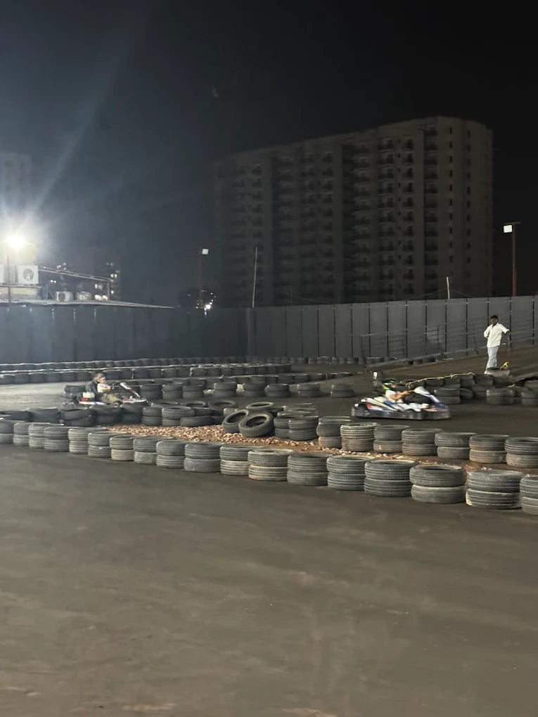 Adults race go-karts at night on a track lined with tires, with a tall building in the background.