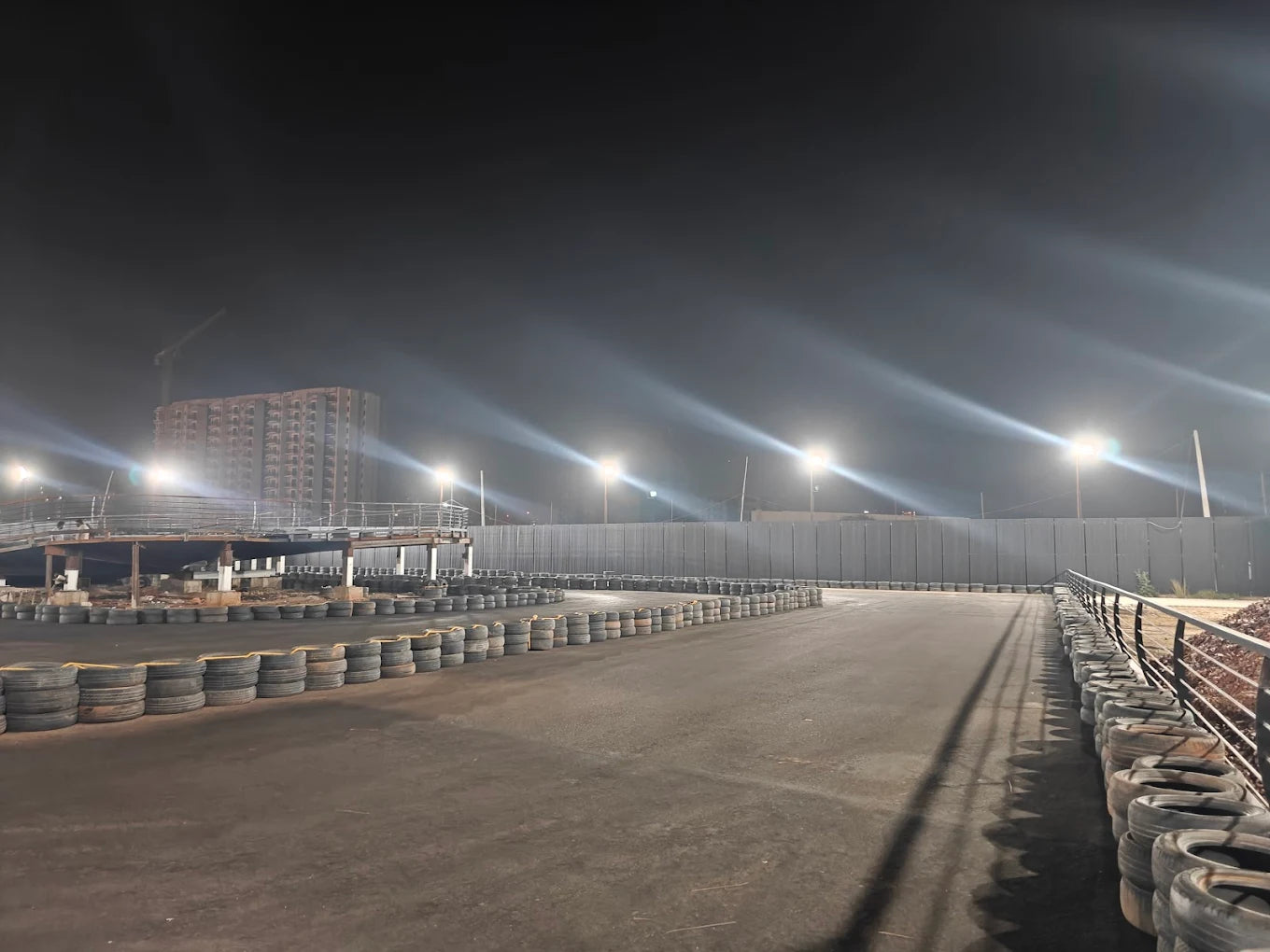 A go-kart track at night, illuminated by bright lights, with a building under construction in the background.