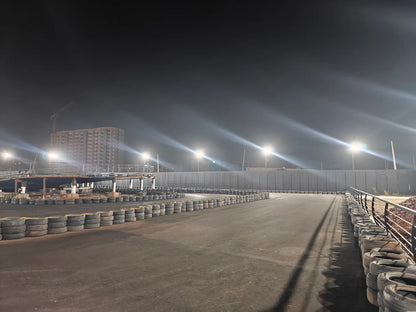 A go-kart track at night, illuminated by bright lights, with a building under construction in the background.