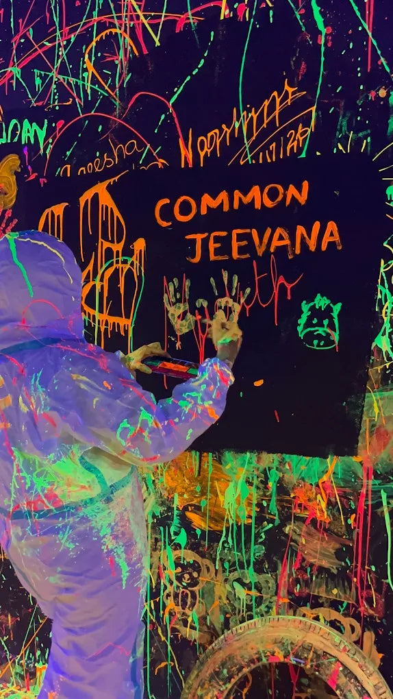 A person in a paint-splattered suit adds handprints to a black canvas covered in neon paint splatters. The canvas has "COMMON JEEVANA" written on it.