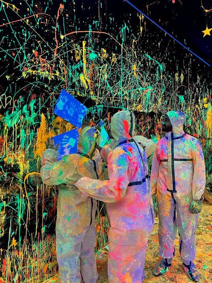 People in protective suits splatter paint on a black wall in a neon-lit room.