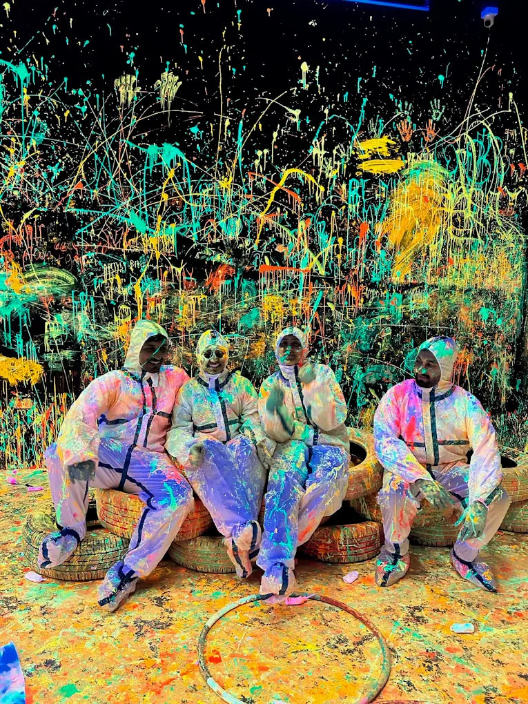 Four adults in protective suits sit on tires in a room splattered with colorful paint.