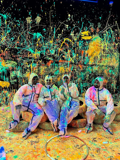 Four adults in protective suits sit on tires in a room splattered with colorful paint.