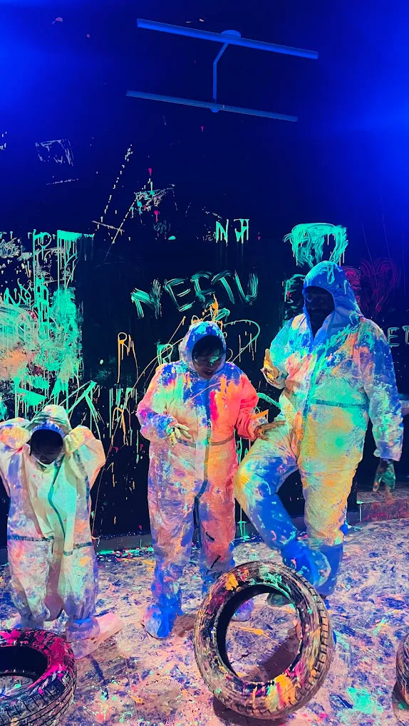 People in paint-splattered suits stand in a dark room with neon graffiti and paint splatters on the walls and floor.