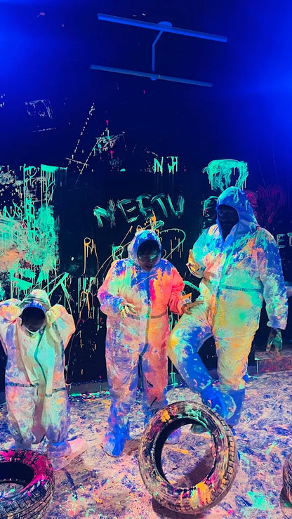 People in paint-splattered suits stand in a dark room with neon graffiti and paint splatters on the walls and floor.