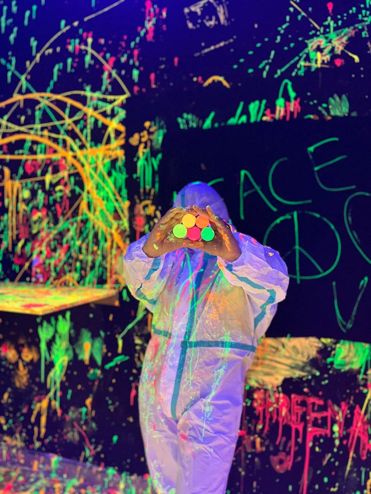 Person in a white suit holds colorful paint pots in a neon-splattered room.