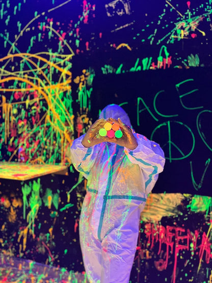 Person in a white suit holds colorful paint pots in a neon-splattered room.