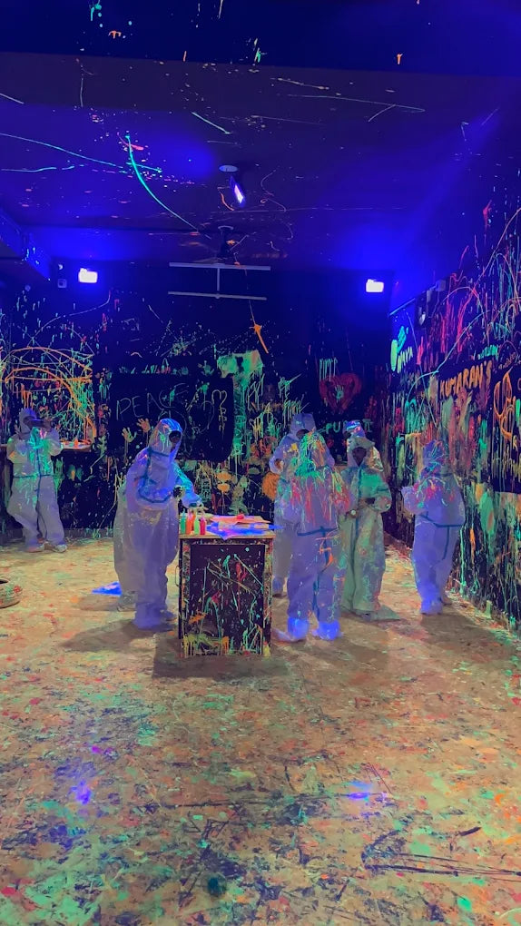 People in protective suits splatter paint in a dark room illuminated by blacklights. The walls and floor are covered in colorful paint splatters.