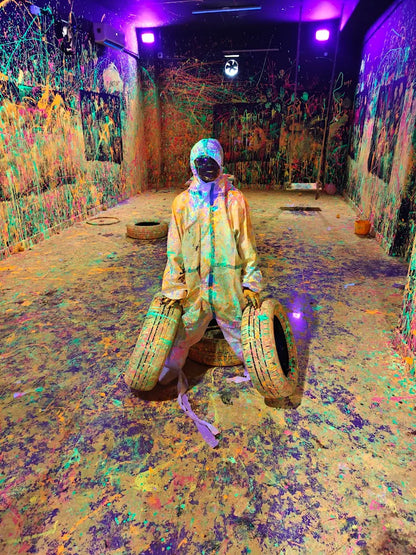 A person in a protective suit sits in a room splattered with neon paint, holding two tires. The room is dark with purple and pink lighting.