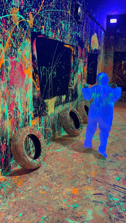 A person in a blue suit stands in a room splattered with neon paint, with tires on the floor.