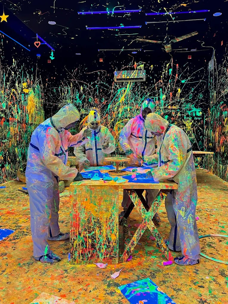 People in protective suits splatter paint in a dark room covered in colorful paint splatters.