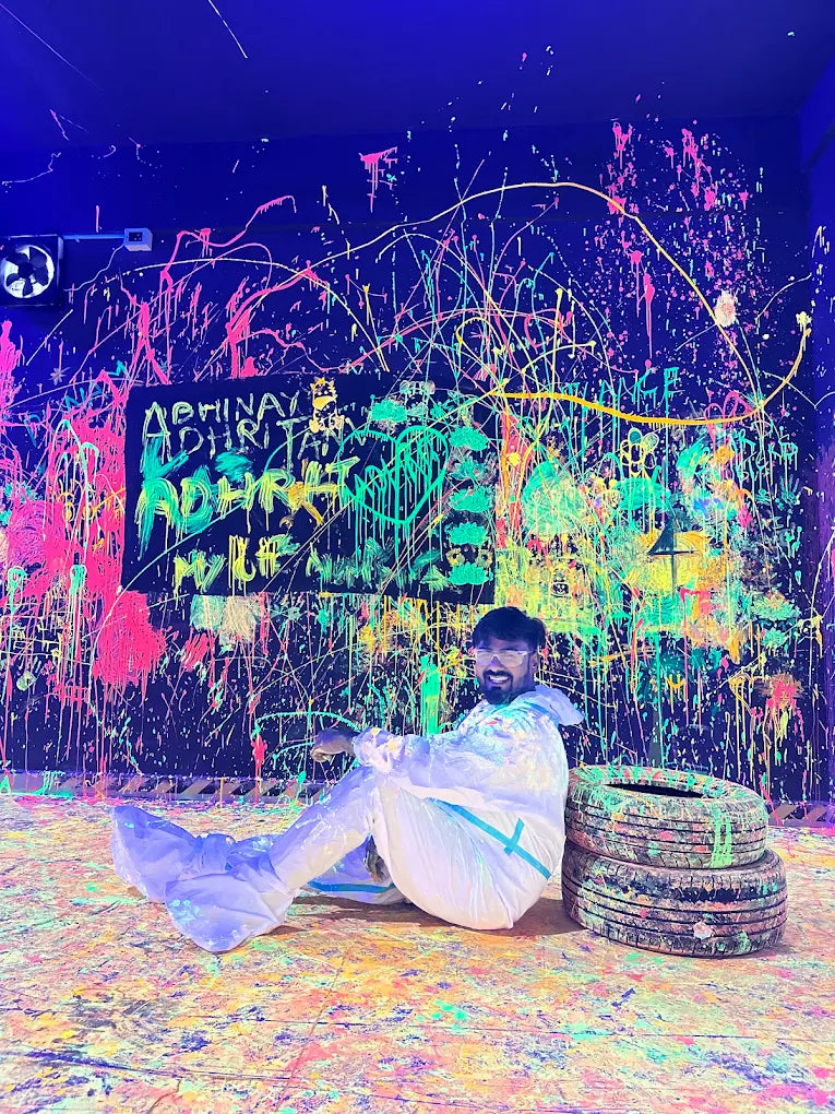 A young man in a white suit sits on a paint-splattered floor in a neon-lit room, with a graffiti-covered wall behind him.