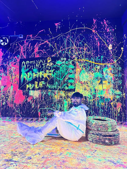 A young man in a white suit sits on a paint-splattered floor in a neon-lit room, with a graffiti-covered wall behind him.