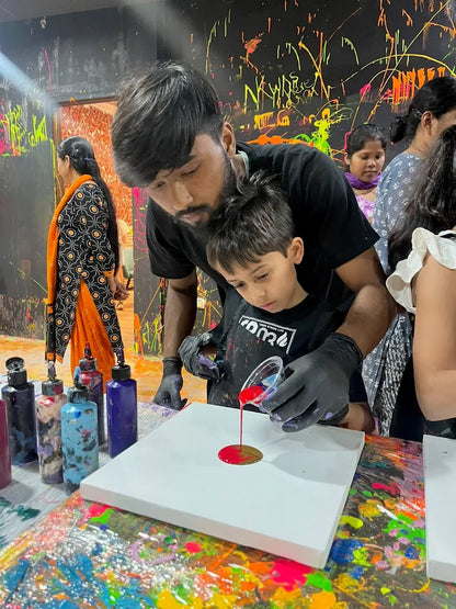 A man and a young boy pour red paint onto a white canvas in a colorful art studio.