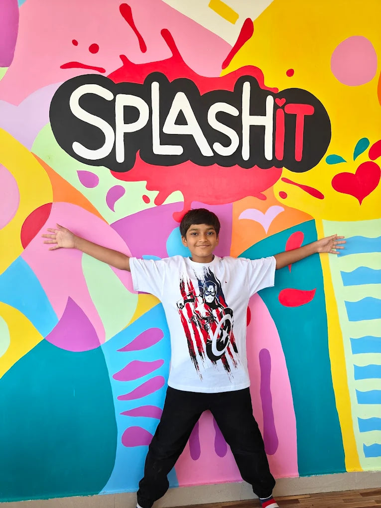 A young boy stands with arms outstretched in front of a colorful mural that says "SPLASH IT".