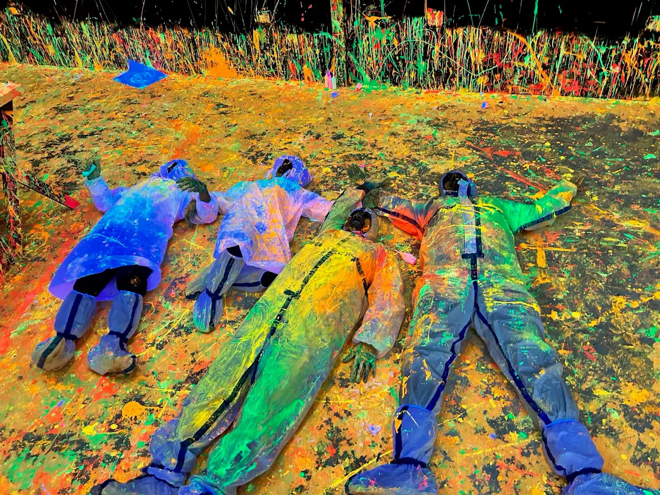 People in protective suits lie on a colorful, paint-splattered ground.