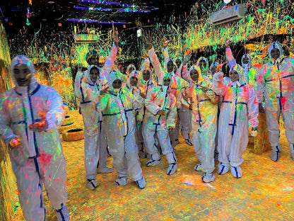 A group of young people in white protective suits are covered in neon paint splatters in a dark room. They are smiling and posing for a photo.